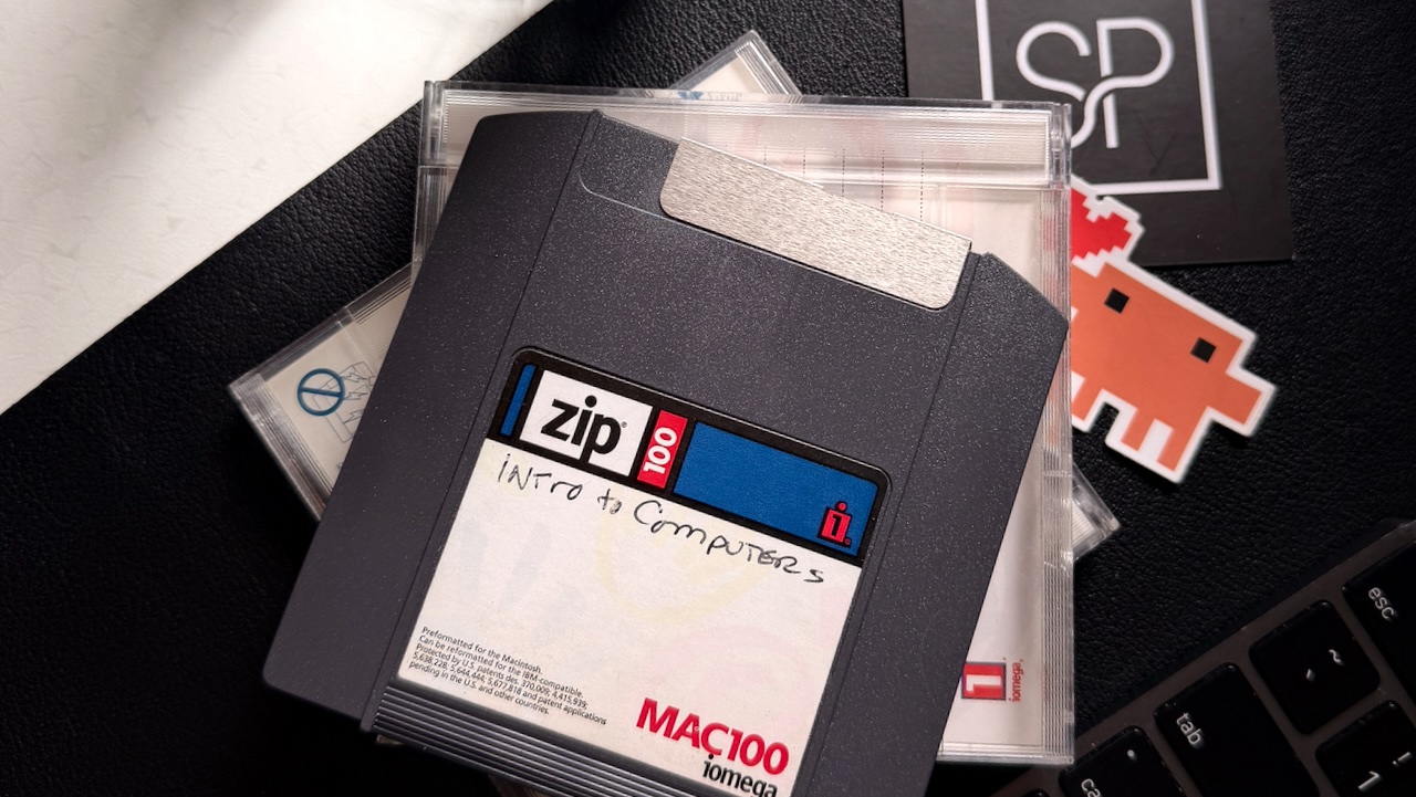 A clear plastic case holding a black Iomega Zip 100 disk hand-labeled "Intro to Computers," resting on a keyboard beside a Studio Practice sticker.